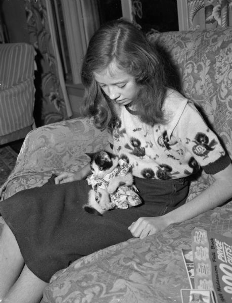 Bubbles, a black and white kitten sitting on the lap of its owner, Janet Kanneberg, eighth grade, 1252 Sherman Avenue. Janet has a wardrobe for Bubbles and her kitten is wearing a colored house frock.