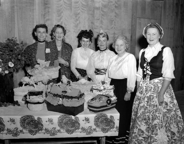 Six women in ethnic dress are shown standing behind decorated lunch baskets.  They are at the first social event held in Our Lady Queen of Peace's new school building in the Westmorland neighborhood. A square dance and basket supper raffle were held in the auditorium. The women shown (left to right) are: Irene Topp, Bernadine Handel, Margaret Denton, Mrs. Schumann, Marie Purcell and Frances Forster.