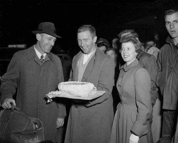 After a 14-6 Badger football victory over Northwestern, a crowd of 3,000 fans jammed the North Western Rail Road Station to welcome the team and present them a cake. Homecoming Queen Carolyn Hunn (right) presents the cake to football team Capt. Robert "Red" Wilson (center) and Coach Ivan "Red" Williamson (left) are looking on.