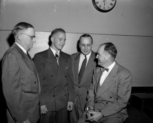 The four main speakers at Madison East High School's 27th annual football banquet stand together beneath a clock. They are (left to right): Foster Randle, principal of East High School; Wendell "Windy" Gulseth, captain of the team; Herbert "Butch" Mueller, coach of the team; and John Walsh, University of Wisconsin boxing coach.