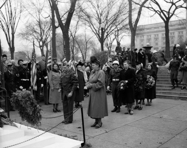 A crowd gathers to participate in a service to observe Armistice Day at the State Street entrance to the Wisconsin State Capitol building. At center left, Clarence L. Skinner, 2025 Sherman Avenue, is saluting after placing a wreath.  Other wreath-bearers are left to right: Mrs John (Mary) Coyne, 13 South Mills Street, American Legion Auxiliary; Mrs. Howard Tholo, 1000 Bowman Avenue, Legion Auxiliary; Mrs. George (Julia) Stahl, 817 Gorham Street, United Spanish War Veterans's Auxiliary; and Patricia McGuine, 250 Division Street, Catholic War Veterans.