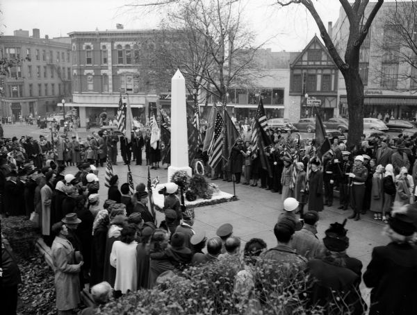A community service to observe Armistice Day at the State Street's entrance to the Wisconsin State Capitol building. Flag bearers and a crowd of people surround a cenotaph with wreaths propped against its base.