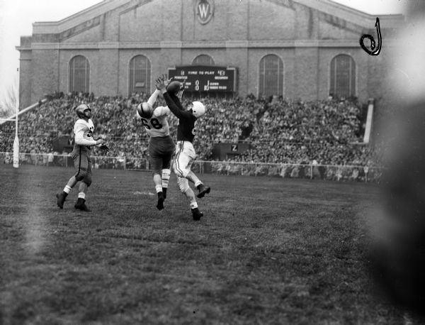University of Wisconsin end Tilden Meyers reaching for a pass in a football game against Iowa at Camp Randall. Iowa defenders are Bill Greene (#68) and Bob Longley (#52). The crowd in front of the Field House is in the background.