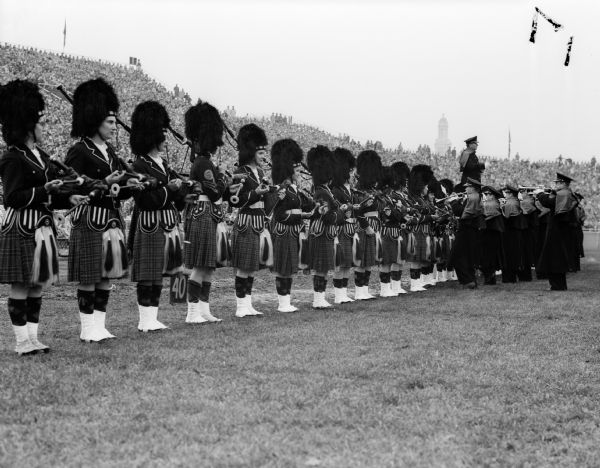 The University of Iowa's Scottish Hilanders marching band wearing kilts and bear fur hats while standing on the sidelines at Camp Randal Stadium.