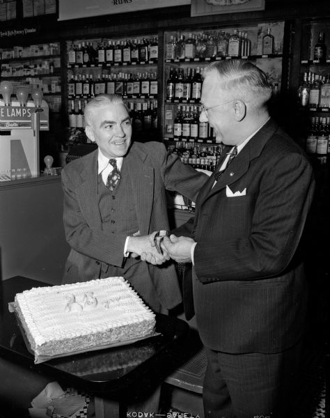 Robert A. Knoll (left) receives a gold watch in honor of his 25 years of service with Walgreen Drug Stores. It is being presented by E.A. Rintelmann. Mr. Knoll is the fountain manager at the 32 East Mifflin Street store. On the table next to them is a cake decorated with the words: "25 yrs."