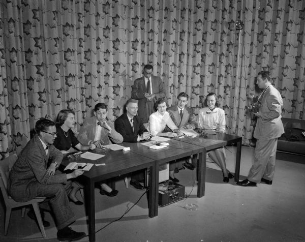 Six University of Wisconsin students and one professor sitting on a panel to record their aspirations for the second half of the twentieth century for "Parade Magazine," a Sunday newspaper supplement. Seated, from left, are Thomas Engelhardt, Wauwatosa; Charm Bolles, Janesville; Karl Meyer, New York; Professor Robert C. Pooley, the round table moderator; Janet Williams, Kenilworth, Illinois; James Cristoph, Waukesha; and Sylvia Fudzinski, West Allis. Standing behind the panel is Sid Ross, "Parade" writer, and at right is his brother, Ben, "Parade" photographer.