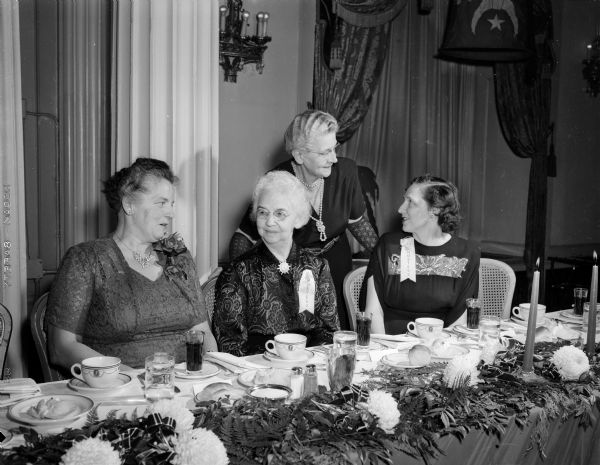 Wives of Zor Shriners sitting beside a decorated table at a banquet held at the Crystal Ballroom at Hotel Loraine. From left at the table are Mrs. Lionel (Helen) Moore, Mrs. Clyde (Ila) Wilson, and Mrs. Edward E. (Inez) Vincent, all of Madison. Standing is Mrs. Alf (Mayme) Peterson, also of Madison.
