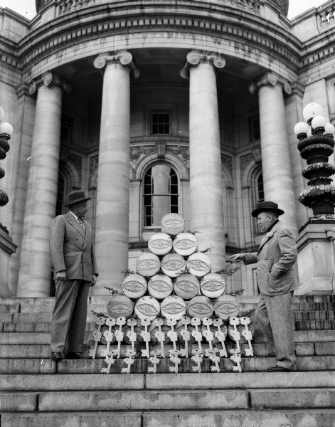 Governor Oscar Rennebohm (left) inspecting the 21 wheels of Marathon County cheddar cheese and keys to Wisconsin's vacation land on the steps of the Wisconsin State Capitol. Joe Mercedes, Governor Rennebohm's good will ambassador, will take the items with him on a tour through Missouri, Illinois, Indiana, Ohio, Kentucky, Tennessee, Georgia, Alabama, Mississippi and Louisiana to promote Wisconsin tourism.