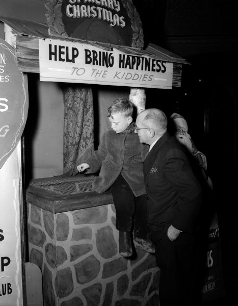 Robert Hoffman dropping coins into the wishing well at the Capitol Theatre for the Empty Stocking Club and the Kiddie Camp fund to "Help Bring Happiness to the Kiddies" while Capitol Theatre manager, Fred Reeth, is looking on.