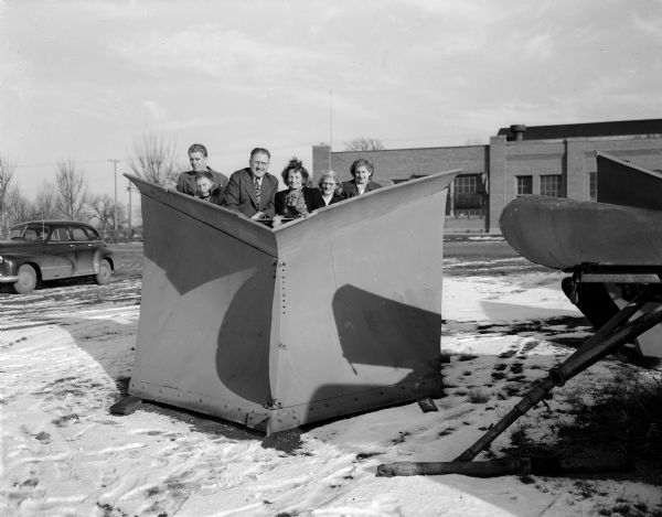 Six men and women from the Frost-Russell family and a dog stand behind a snow plow blade for a group portrait.