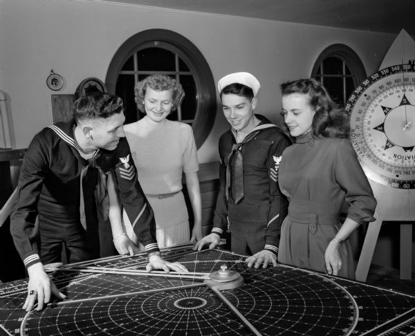 Naval Reserve sailors Ralph Bakken (left), and Richard J. Nelson show a ship's maneuvering or plotting board to Irene Strand, second from the left, and Kay Franke at the United States Naval Reserve Surface Division 9-212 surprise party. The party was given by the officers for the enlisted men at the new training center, located at 1046 East Washington Avenue. The training center has a complete duplication of the bridge of a ship.
