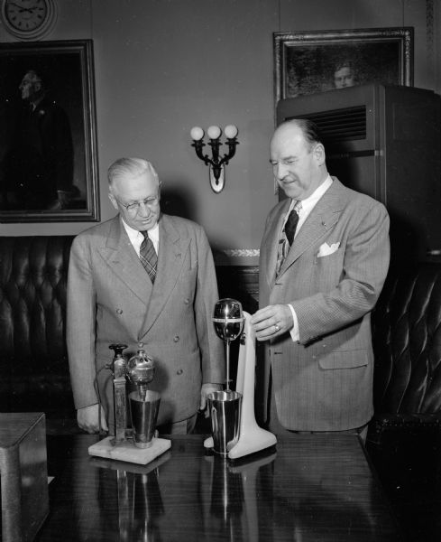 Governor Oscar Rennebohm (right) stands with an unidentified man in the Governor's office while looking at two Hamilton Beach soda mixers on a table. The Hamilton Beach company is located in Racine, Wisconsin.