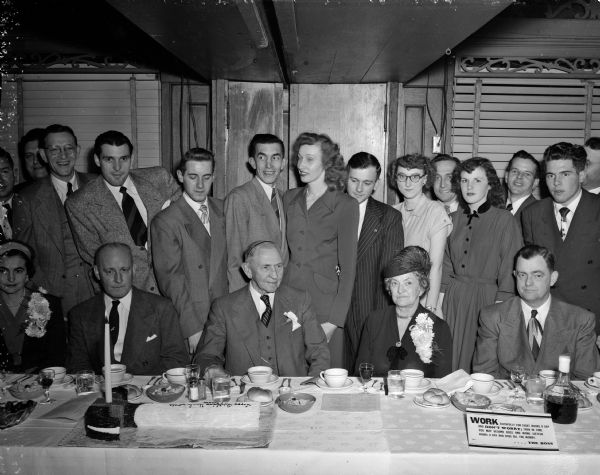A group gathers for a portrait around a table at the Park Hotel during the birthday dinner of Madison businessman Louis Hirsig. Seated at the dinner table are, left to right: Guy Martin, a son-in-law; Louis Hirsig; Mrs. (Marie) Hirsig; and another son-in-law, Frank Doyle. Standing behind them are many of the employees of Wolff, Kubly, and Hirsig Company.