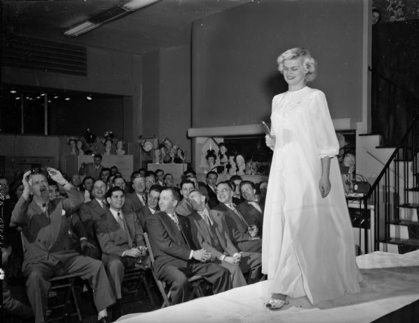 A woman modeling a peignoir and matching night gown for a crowd of men during Carmen's Style Show for Men. Roundy Coughlin is seen in the front row cleaning his glasses.