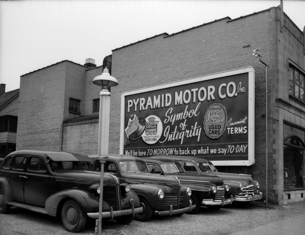 Four cars park in the Pyramid Motor Company used car lot at 721 University Avenue. A company billboard is on the side of the Dictaphone Sales Agency building at 731 University Avenue.