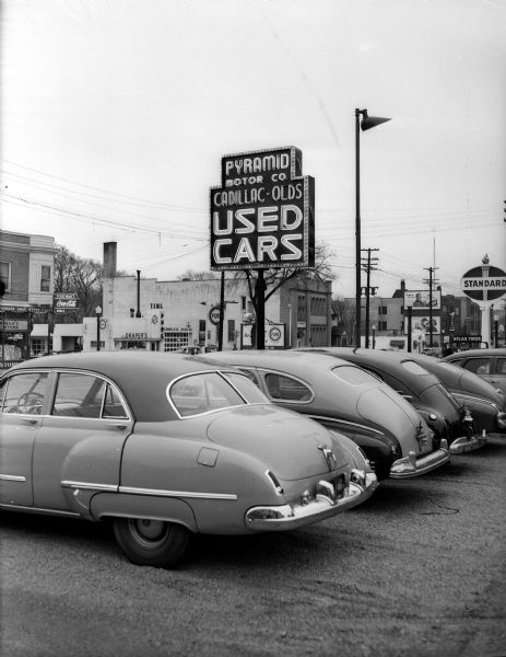 Cars park in the Pyramid Motor Company used car lot at 721 University Avenue.  The Standard Oil Company service station sign can be seen in the background at 701 University Avenue. Businesses across the street include Tiedeman Drug Store (702-704), Drapers Pure Oil service station (630-632), and Fitch-Lawrence Funeral Directors (626).
