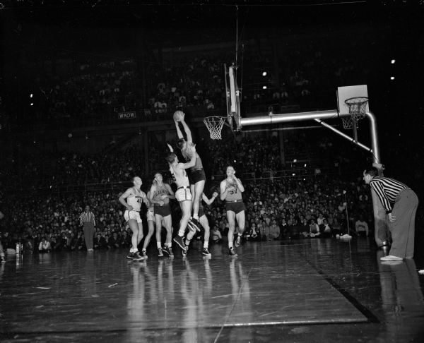 An action photograph of a basketball game shows an exciting moment in the competition. Illinois' Burdette Thurbly (#14) is going up high to take a rebound from Wisconsin's "Danny" Markham (#38). Other players in the background are Wisconsin's Fred Schneider (#40), and Illinois team members Roy Gatewood (#8), and Rod Fletcher (#37).