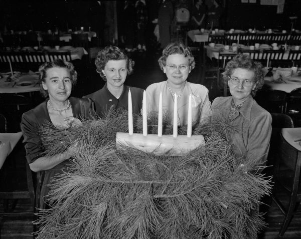 Committee members for the Nakoma 12th Night Party gather around a table set with candles and evergreen branches in a banquet room.  They are, from left to right, Mrs. John F. Stauffer, 4113 Hiawatha Drive; Geraldine LaBonne, 4105 Cherokee Drive; Leeta Howard, 4154 Nakoma Road; Elizabeth Omernik, 4125 Nakoma Road.