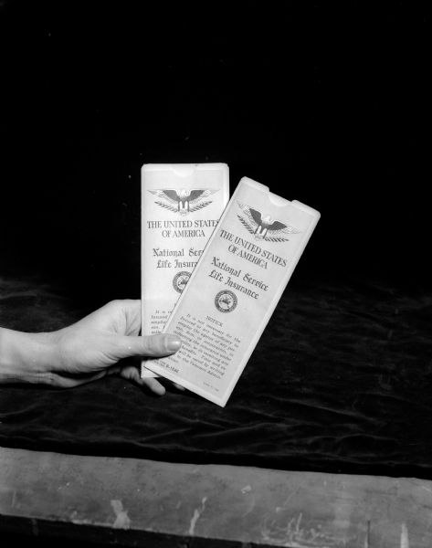 A person's hands holding two U.S.A. National Service Life Insurance Policies. About $60,000,000 in GI Insurance dividends will be divided among 347,000 Wisconsin veterans and next-of-kin. Note: One of four images used in a montage of Don Dickinson, World War II veteran, and his daughter Linda Sue.