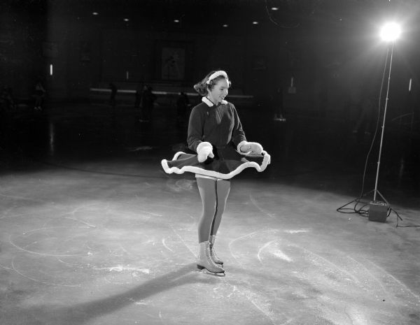 Dorothy Bell, a member of the Madison Skating Club's Junior Club, is shown spinning while wearing a skirted skating costume at their indoor rink at Truax Field. The original caption mentions the photographer used strobe lights to "freeze" the skater's spinning skirt. The negative shows a strobe light on a tall stand on the right side.