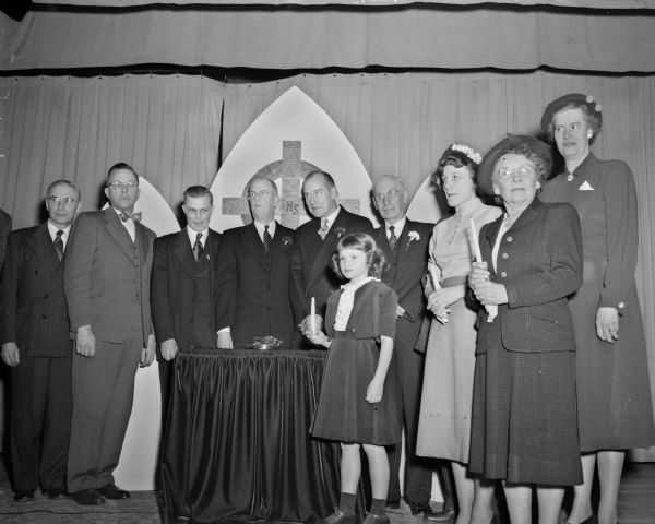 Group portrait of the principal participants in a mortgage burning ceremony at First Congregational Church, 1609 University Avenue. The congregation is celebrating the 110th year of their existence and 20 year anniversary of their church building. Left to right: Lea Gunderson; A.G. Gehner; R.W. Sackett; the Rev. Alfred Swan, pastor;  the Rev. Robbins W. Barstow, former pastor; Clayton Haswell; Mrs. John E. (Doris) Anderson; Mrs. L.W. (Bess) Bridgman; and Mrs. Fred E. (Elizabeth) Risser. In front of the group is Christine, daughter of John E. and Doris Anderson.