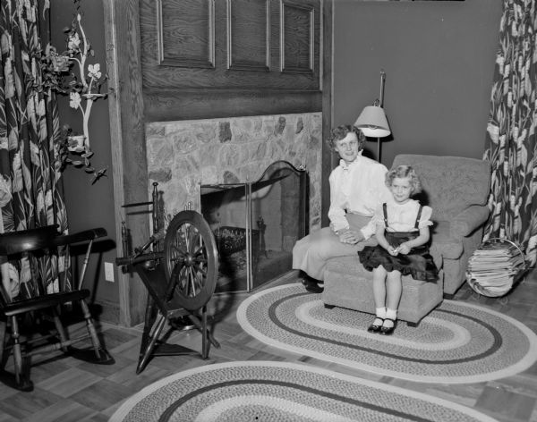 Living room of the residence of Mr. and Mrs. Bernard F. (Sylvia) Killian, 3814 Hillcrest Drive. Mrs. Killian and her daughter Elaine are shown seated in front of the fieldstone fireplace with a spinning wheel in front of them. Mr. Killian, a Madison builder who has his office in the house, designed the family home to suit the location and to fill the owners' needs.