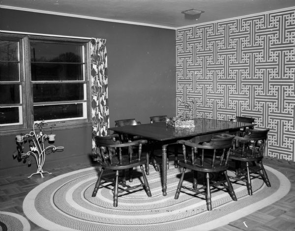 Dining "L" of the residence of Mr. and Mrs. Bernard F. (Sylvia) Killian, 3814 Hillcrest Drive, featuring a geometic patterned paper on one wall that uses the colors of the room's decor: forest green, chartreuse, cherry, and cocoa. Draperies on either side of the large picture window also use the same colors as does the oval braided rug under the cherry dining room set.