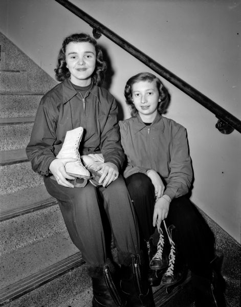 Two high school girls with ice skates on a staircase at their school as they prepare to leave for the nearest skating rink. Pictured left to right are Mary Jean Whalen and Kay Aspel.