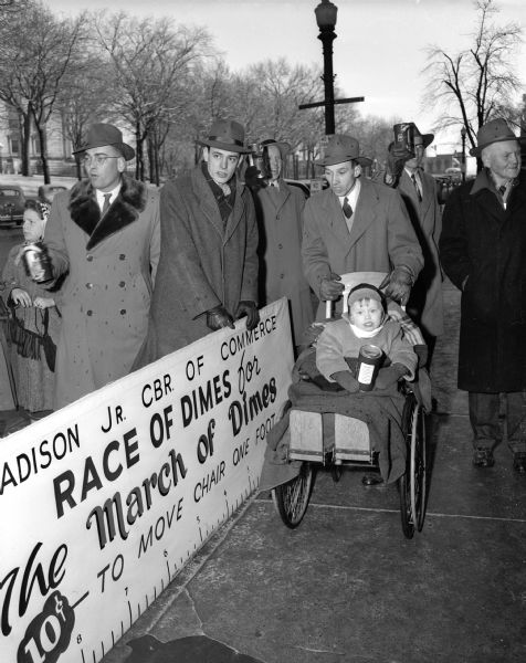 Four-year-old Paul Rosen, son of Mr. and Mrs. Paul R. (Agnes) Rosen, 118 Jackson Street, is all smiles at the Junior Chamber of Commerce March of Dimes Wheelchair Race. Pictured with Paul, from left to right, are: John Jenswold, Chairman of the race, Hank Bush Jr., Gene Erickson, and Robert Pride, standing behind Paul's wheelchair.