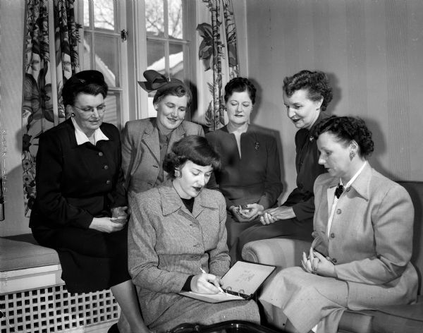 Six Alpha Gamma Delta alumnae members assist with arrangements for a benefit bridge party.  Seated in the foreground are: Mrs. H.L. (Esther) Manthei, 44306 Doncaster Drive, left and Mrs. Robert M. (Hazel) Wheeler, 2441 Fox Avenue. In the background, left to right:  Mrs. J.A. (Helen) Keenan, 2633 Chamberlain Avenue; Mrs. Harrison A. (Kath) Smith, Jr., Maple Bluff; Mrs. Milton J. (Edith) Heisman, 432 North Frances Street; and Dr. Madeline Thornton, Edgewater Hotel.