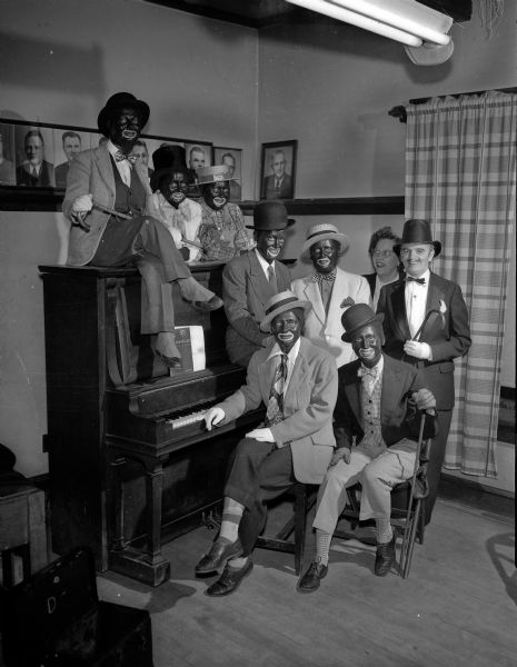 Nine women, all but two in blackface and all dressed in drag for a minstrel show, presented by a St. Bernard's Catholic Church Christian Mothers group. Seated on the piano is Mrs. Frank (Clara) Zeier; standing behind the piano, left to right: Mrs. Joseph (Lucille) Nolter and Mrs. A.L. (Eleanore) Derra; standing in the middle row, left to right: Mrs. Carl (Doris) Blum, Mrs. J.E. (Alice) Kleiner, Mrs. John (Marie) Schroeder, director; and Mrs. Nicholas (Flora) Raffay; seated in the foreground: Mrs. William (Helen) Rethke and Mrs. Wallace (Erma) Heisig.