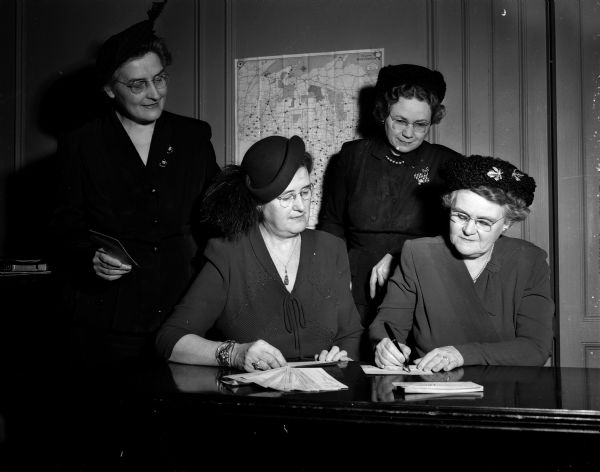 Three members of the Madison Council of Church Women observe Mrs. Leonard (Fay) Howell, wife of the city manger, as she signs the first card of the Eumenical Register, a project to put a million American protestant women on record for Christian unity throughout the world. Standing are, left to right:  Mrs. O.I. (Marie) Dhein, state president of the Wisconsin Council of Church Women; and Mrs. Leon W. (Mary) Petersen, president of the Madison council. Seated: Fay Howell and Mrs. Cyril (Leeta) Howard, chair of the council's key women.