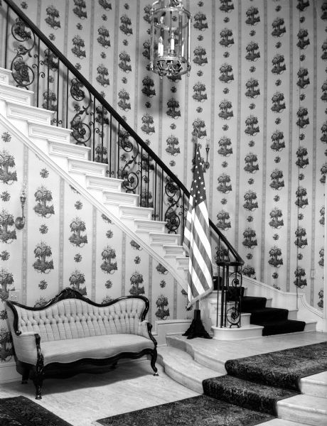Reception hall at the Governor's residence, 101 Cambridge Road. A curving staircase rises above the reception hall near the entry door to the Wisconsin governor's new executive residence. Period wallpaper in gray and pastels was Governor's wife Mary Rennebohm's first step in redecorating the house.  An antique settee is one of many pieces of furniture brought from the old executive residence.
