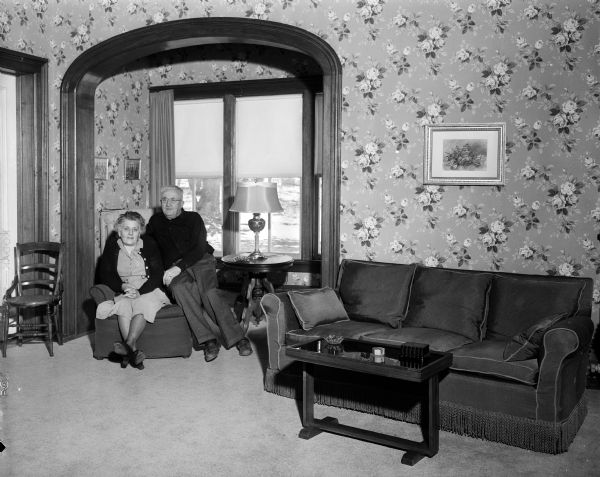 One of the two living rooms of the home of Mr. and Mrs. H.D. Brainard, the former 1878 "old Don Johnson farm" in Bear Valley, which the Brainard's purchased after the last member of the Johnson family died and willed the farm to the Salvation Army. Mr. and Mrs. Brainard are pictured seated in their living room decorated with tan and green floral wallpaper, gray carpeting, a deep maroon ceiling, and family antiques.