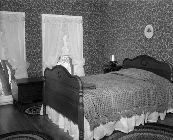 A bed and wooden chest stand in the master bedroom of the home of Mr. and Mrs. H.D. Brainard. The home was the former 1878 "old Don Johnson farm" in Bear Valley, which the Brainard's purchased after the last member of the Johnson family died and willed the farm to the Salvation Army. Fifty-year old wallpaper featuring pinky rose flowers on a cream background and a rose painted ceiling provide the color theme for the room.
