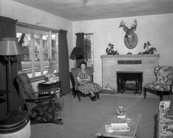 The living room of the home of Mr. and Mrs. Scholl on East Montroe Street. Mrs. Scholl is seated in a lipstick red chair next to the Lannon stone fireplace. In addition to lipstick red, the colors used in the room's decor are old gold, forest green, and gray.