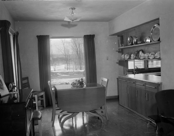 The decor of the  dining room "L" in the home of Mr. and Mrs. Clarence Scholl, located on East Monroe Street, repeats the colors used in the living room of the home: forest green, lipstick red, old gold, and gray. Built-in shelves along one wall with a cut-out area below link the dining room with the kitchen.