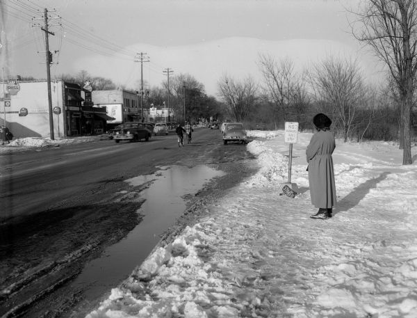 A woman is standing at a bus stop at the end of Monroe Street, the entrance to Nakoma, where puddles from melting snow cause water from passing traffic to splash bus riders waiting at the stop. Businesses that can be seen across the street include the F & D Grocery, 3518 Monroe Street; Elver's Shell Service Station, 3418 Monroe Street; and Mallatt Pharmacy, 3410 Monroe Street.