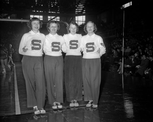St. Croix Falls cheerleaders stand along the sidelines in the University of Wisconsin fieldhouse at a high school basketball tournament. They are, from left to right: Carol Lau, Betty Olson, Judy Greenquist, and LaDonna Peterson.