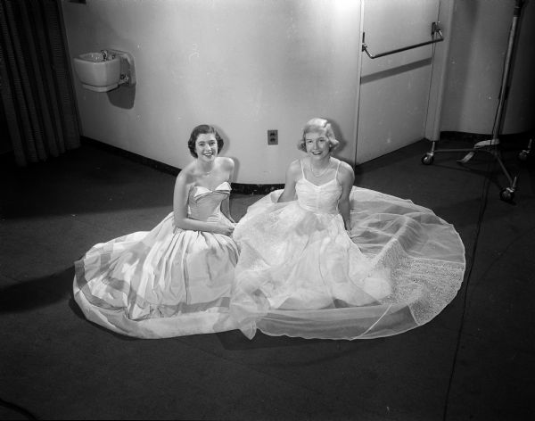 Two female members of the Court of Honor for the University of Wisconsin military ball sitting on the floor wearing formal gowns. At left is Joan Wolf of Highland Park, Illinois, Alpha Epsilon Phi member. Barbara Becker, Kappa Kappa Gamma, of Wauwatosa, Wisconsin is sitting beside her. A bubbler (water fountain) is on the wall in the background.