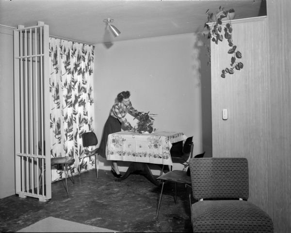 A woman decorates a modern dining-room. At right is a large movable wardrobe that the home owner may use as an adjustable room divider as the family needs change. The home builders were Ivan Gregory and Jack Klund of Space, Inc.  They built this house at (probably) 5110 Shore Acres Road as well as other houses on Shore Acres Road.