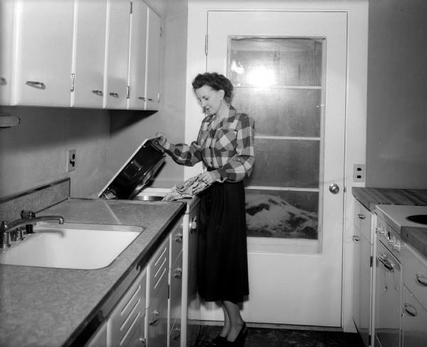 A woman is standing in a modern kitchen while peering into a top-loading dishwasher. The home builders were Ivan Gregory and Jack Klund of Space, Inc.  They built this house at (probably) 5110 Shore Acres Road, as well as other houses on Shore Acres Road.
