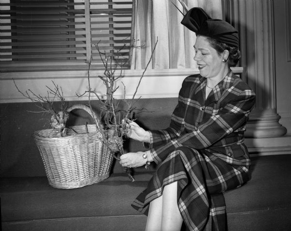 A woman wearing a dress and hat, probably June D. McLean, arranges flowers in a wicker basket while seated on a couch.
