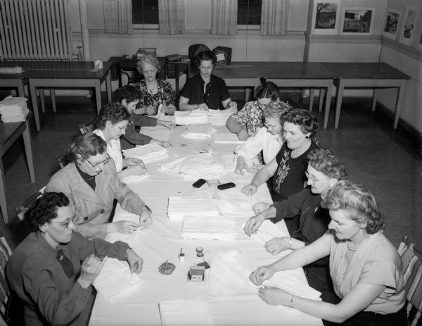 Members of the Eagles' auxiliary making dressings for cancer patients while sitting around a table at the Woman's Building. Around the table from the left are Carol France, Florence McCann, Helen Kohl, Olga Kreil, Lydia Wolfe, Jo Newman, Lillie Becker, Stella Neby, Margaret Dolva, Elsie Ewing, and Adeline Dettner.