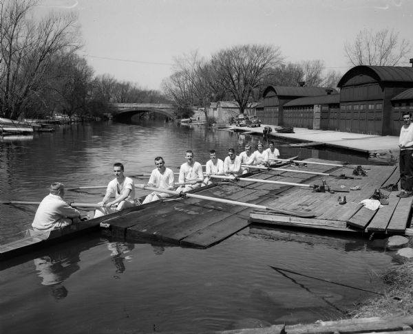 A University of Wisconsin rowing crew prepares for a workout in the Yahara River aboard a scull. Trachte metal boat houses line the shore at right, and the Johnson Street bridge is visible in the background.
