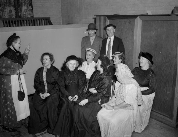 A group of Westminster Presbyterian Church members in old fashioned dress recreate an old-time singing school as a fund raiser to bring five displaced persons from Europe to Madison. Mrs. Lawrence E. (Vinnie) Blair, left, leads the singers. Front row, left to right: Mrs. Wallen C. Laura) Klein, Mrs. H.L. (Jean) Jones, Mrs Richard E. (Eleanor) Pritchard, and Mrs. Carolin McNelly.  Second row: Mrs. Lionel G. (Helen) Moore, Mrs. Charles W. Lucille) Oakey, Jr, and Mrs. Chellis Botts.  Back row: Wallen C. Klein and H.I. Jones.
