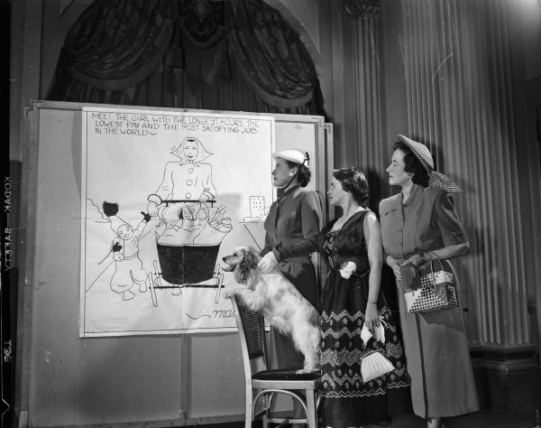 Three of the models participating in the Jaycettes' fashion show admire a large-sized cartoon that will be used in the show, inspired by State Journal cartoonist Dorothy Bond's "Mama." Left to right: Mrs. Jerry (Patricia) Butler, 745 University Avenue; Mrs. Wade (Aleen) Plater, 403 Farwell Drive; and Mrs. D.J. (Florence) Leigh, 2146 Lakeland Avenue. Also pictured is a Cocker Spaniel, Schmoo, who will appear in the show as well.