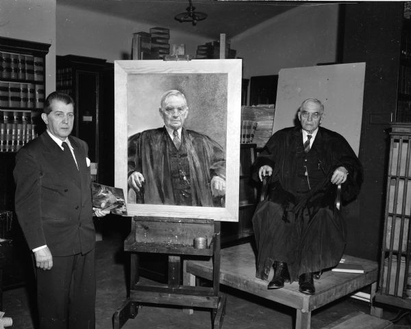 Noted Chicago artist Christian Abrahamson holds a palette and paintbrushes as he stands next to a portrait of the retired chief justice of the Wisconsin Supreme Court Marvin B. Rosenberry. The chief justice sits in the chair where he posed for the painting, behind the easel on an elevated platform.