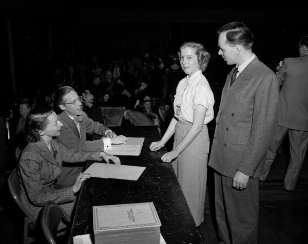 Badger Spelling Bee champion Arlene Brown stands before seated judges Mrs. Robert Taylor and Morton Schroeder, as Master Pronouncer Paul W. Gauger looks on. An audience is in the background.