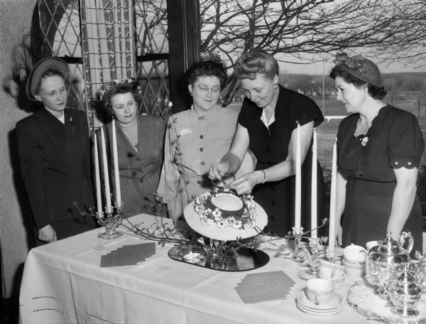 Five women of the planning committee for the Nakoma Golf Club's women's spring tea and style show gather around a decorated table.  Shown at the serving table are, left to right: Mrs. W.H. (Florene) Wendt, social chairman; Mrs. K.O. (Lucille) Nedrebo, publicity chairman; Mrs. J.L. (Mildred)Johnson, music chairman; Mrs. F.D. Chamberlin, table arrangements chairman; and Mrs. John J. (Edith) Drives, president of the Nakoma Women's Organization.